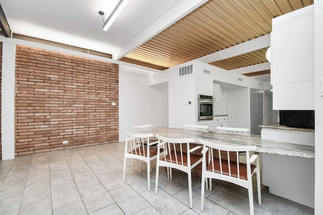 kitchen with brick wall, oven, white cabinetry, beamed ceiling, and light tile patterned floors