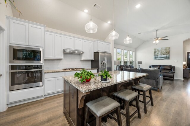 kitchen featuring appliances with stainless steel finishes, under cabinet range hood, vaulted ceiling, tasteful backsplash, and a kitchen breakfast bar