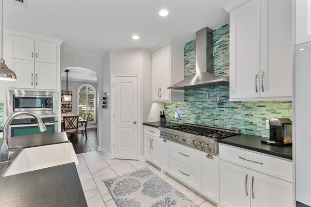 6203 Aspen Pass Dr, Kingwood, TX 77345 - kitchen featuring wall chimney range hood, stainless steel appliances, arched walkways, dark countertops, and white cabinetry