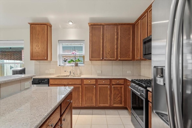 kitchen featuring appliances with stainless steel finishes, a sink, backsplash, light stone countertops, and brown cabinets