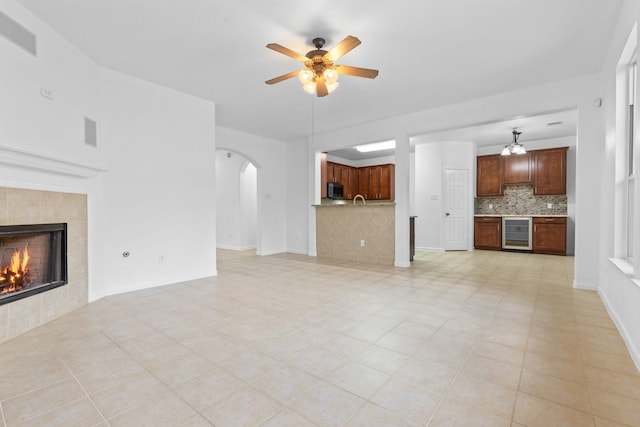unfurnished living room with beverage cooler, arched walkways, ceiling fan, a tiled fireplace, and light tile patterned floors