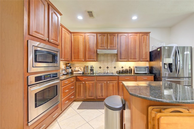 2127 Linden Rock Dr, Katy, TX 77494 - kitchen with stainless steel appliances, dark stone countertops, light tile patterned floors, tasteful backsplash, and brown cabinetry