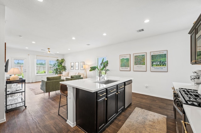 kitchen featuring dishwasher, gas stove, dark cabinetry, a center island with sink, and light countertops