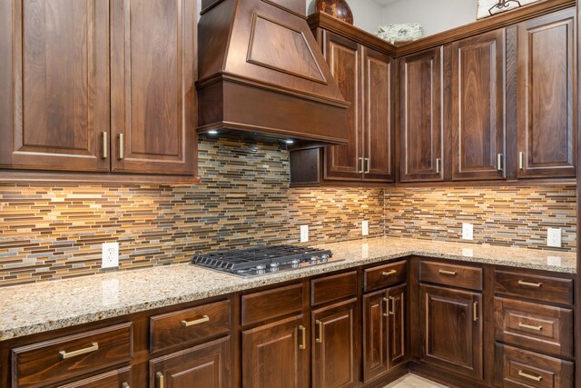 13909 Morgan Bay Dr, Pearland, TX 77584 - kitchen featuring custom range hood, stainless steel gas stovetop, decorative backsplash, dark brown cabinetry, and light stone countertops