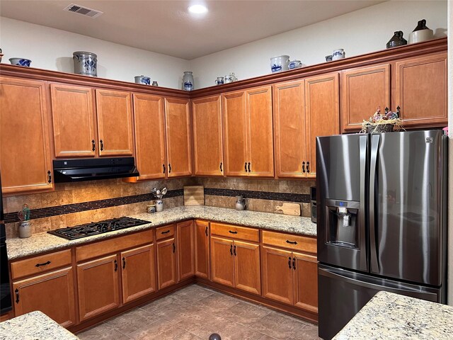 115 Carriage Ct, Magnolia, TX 77354 - kitchen featuring stainless steel fridge with ice dispenser, black gas stovetop, under cabinet range hood, brown cabinetry, and light stone countertops