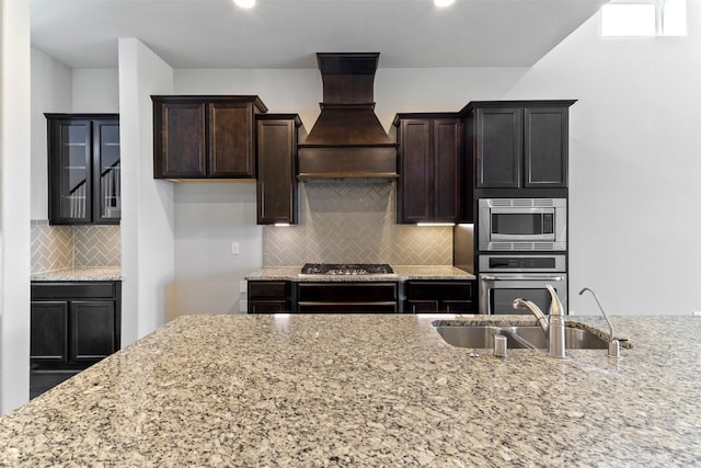 4926 Thunder Creek Ln, Sugar Land, TX 77479 - kitchen with stainless steel appliances, custom exhaust hood, a sink, light stone counters, and dark brown cabinetry