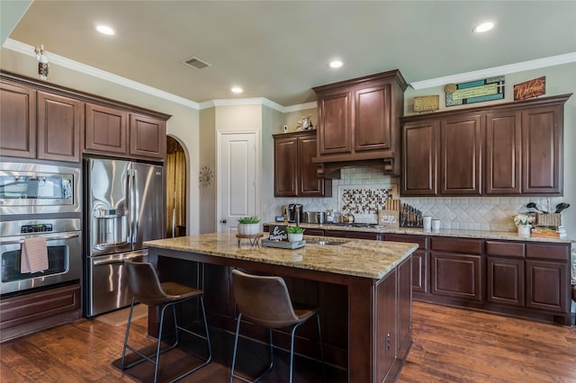10040 Easton Sky Ln, Cypress, TX 77433 - kitchen featuring appliances with stainless steel finishes, a kitchen island with sink, light stone countertops, dark brown cabinetry, and dark wood-type flooring