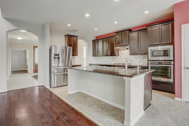 5402 Bending Branch Dr, Missouri City, TX 77459 - kitchen featuring appliances with stainless steel finishes, arched walkways, dark stone countertops, a kitchen island with sink, and under cabinet range hood