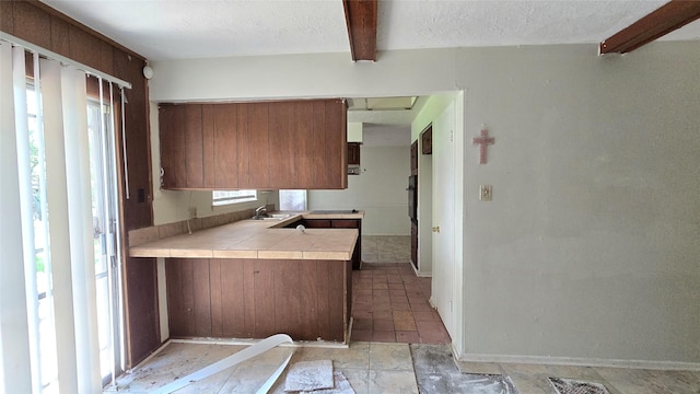 3823 Regency Dr, Houston, TX 77045 - kitchen featuring tile counters, a sink, beamed ceiling, a peninsula, and brown cabinetry