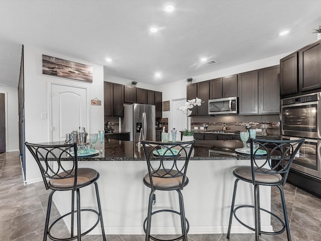 14306 Cedar Pine Ct, Houston, TX 77068 - kitchen featuring appliances with stainless steel finishes, dark brown cabinets, a kitchen bar, recessed lighting, and dark stone counters