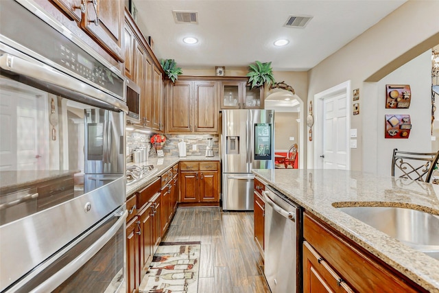 8215 Frontenac Dr, Houston, TX 77071 - kitchen featuring stainless steel appliances, brown cabinetry, decorative backsplash, light stone counters, and arched walkways