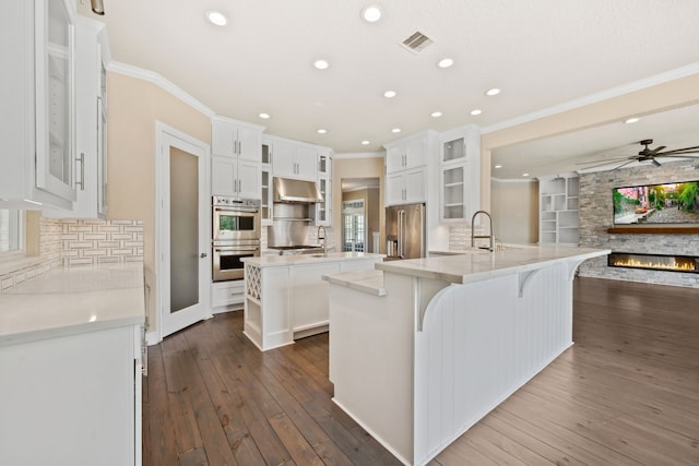 806 Quintana Roo Pl, Seabrook, TX 77586 - kitchen with appliances with stainless steel finishes, a center island with sink, under cabinet range hood, crown molding, and a stone fireplace