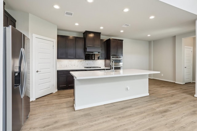 29211 Pikes Peak Dr, Katy, TX 77494 - kitchen featuring appliances with stainless steel finishes, an island with sink, light wood-style floors, backsplash, and light countertops