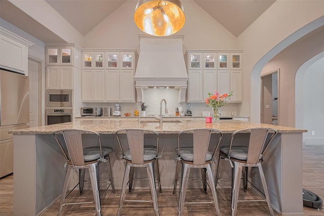 13707 Rivendell Crest Ln, Cypress, TX 77429 - kitchen with appliances with stainless steel finishes, custom exhaust hood, lofted ceiling, a center island with sink, and white cabinetry