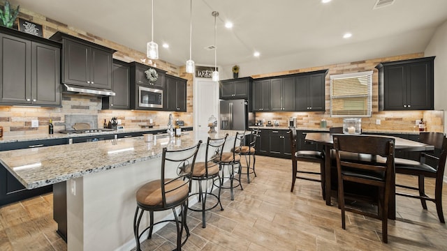 5903 Red River Dr, Manvel, TX 77578 - kitchen featuring stainless steel appliances, under cabinet range hood, a kitchen bar, tasteful backsplash, and light stone countertops