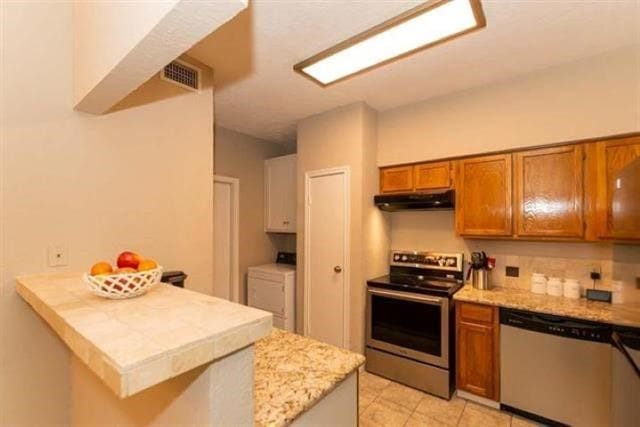 1620 Wellington Pl Unit 303, Beaumont, TX 77706 - kitchen with stainless steel appliances, under cabinet range hood, brown cabinetry, light countertops, and washer and dryer