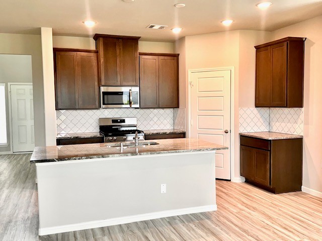 5918 Bright Keel Dr, Katy, TX 77493 - kitchen with stainless steel appliances, a center island with sink, light stone counters, light wood-style flooring, and tasteful backsplash