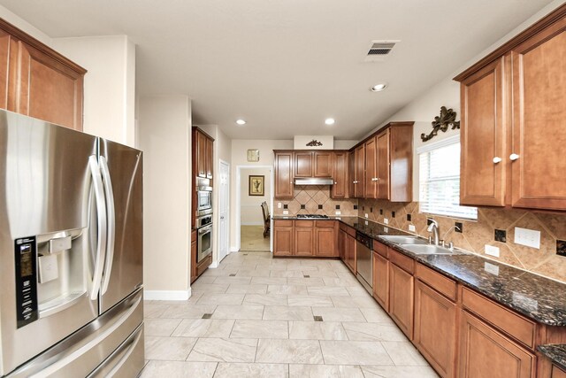 20718 Mansfield Bay Ln, Richmond, TX 77407 - kitchen featuring appliances with stainless steel finishes, brown cabinetry, under cabinet range hood, backsplash, and dark stone countertops