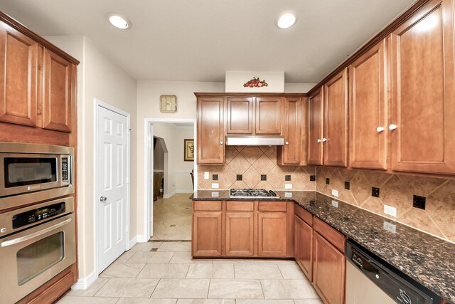 20718 Mansfield Bay Ln, Richmond, TX 77407 - kitchen with appliances with stainless steel finishes, under cabinet range hood, dark stone counters, brown cabinetry, and tasteful backsplash