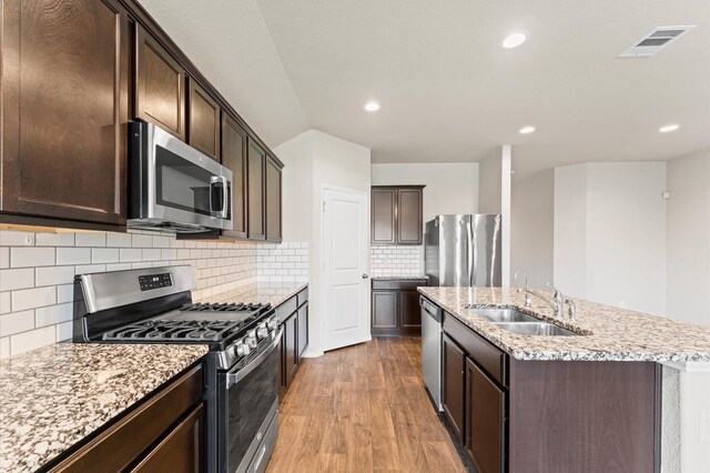 919 Bright Lotus Ln, Rosharon, TX 77583 - kitchen featuring appliances with stainless steel finishes, an island with sink, wood finished floors, dark brown cabinetry, and light stone countertops