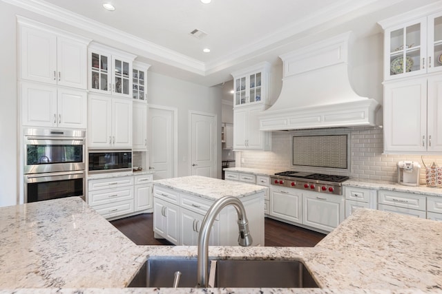 2 N Badger Lodge Cir, The Woodlands, TX 77389 - kitchen featuring appliances with stainless steel finishes, custom exhaust hood, white cabinetry, ornamental molding, and decorative backsplash