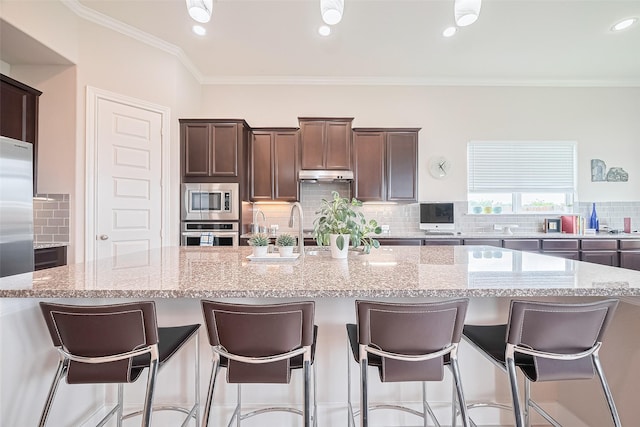 25603 Kearsley Dr, Katy, TX 77494 - kitchen with stainless steel appliances, an island with sink, decorative backsplash, dark brown cabinetry, and crown molding
