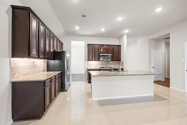 5107 Preserve Park Dr, Spring, TX 77389 - kitchen with appliances with stainless steel finishes, dark brown cabinetry, tasteful backsplash, light stone counters, and a center island with sink