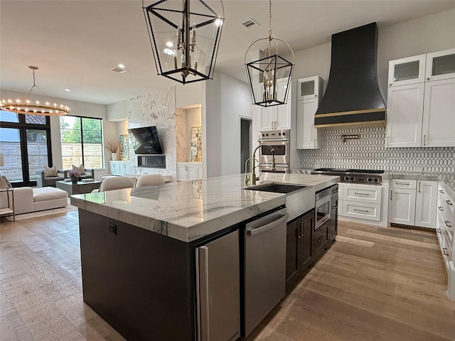 4622 Banning Dr, Houston, TX 77027 - kitchen with custom exhaust hood, appliances with stainless steel finishes, an island with sink, a chandelier, and white cabinetry