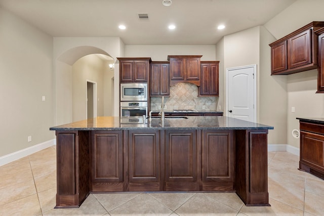 1215 Oxbow Crossing Ln, Rosenberg, TX 77471 - kitchen featuring stainless steel appliances, arched walkways, decorative backsplash, dark stone countertops, and an island with sink
