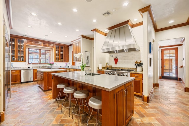 603 W Friar Tuck Ln, Houston, TX 77024 - kitchen with appliances with stainless steel finishes, an island with sink, brown cabinets, crown molding, and wall chimney exhaust hood