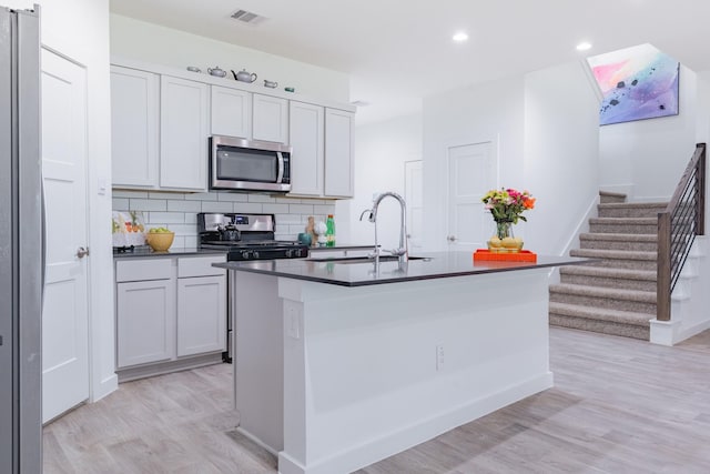 14205 Wonder Lake Ln, Conroe, TX 77384 - kitchen with appliances with stainless steel finishes, dark countertops, light wood-type flooring, an island with sink, and tasteful backsplash