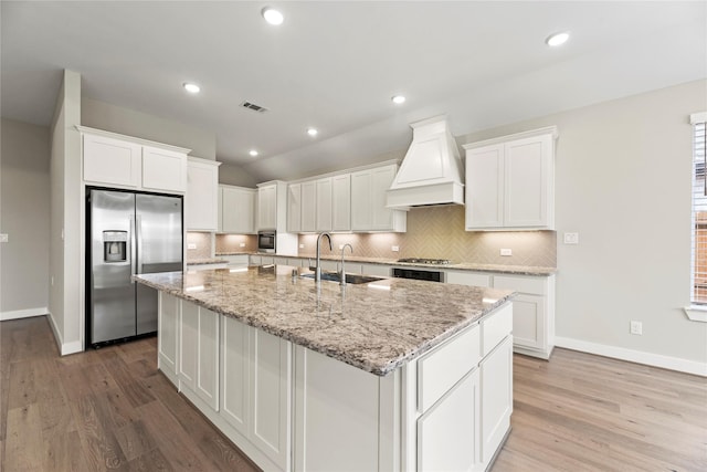 9418 Whispering Bald Cypress Ct, Cypress, TX 77433 - kitchen featuring appliances with stainless steel finishes, custom range hood, a kitchen island with sink, light wood finished floors, and white cabinetry