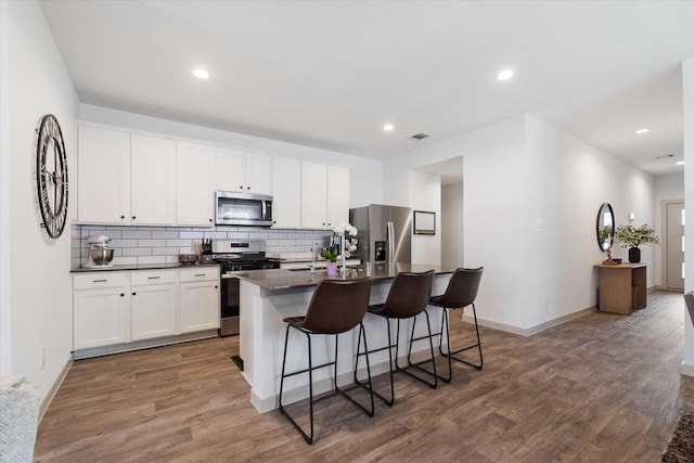 14906 Rain Tree Dr, Baytown, TX 77523 - kitchen with appliances with stainless steel finishes, dark countertops, a center island with sink, decorative backsplash, and wood finished floors