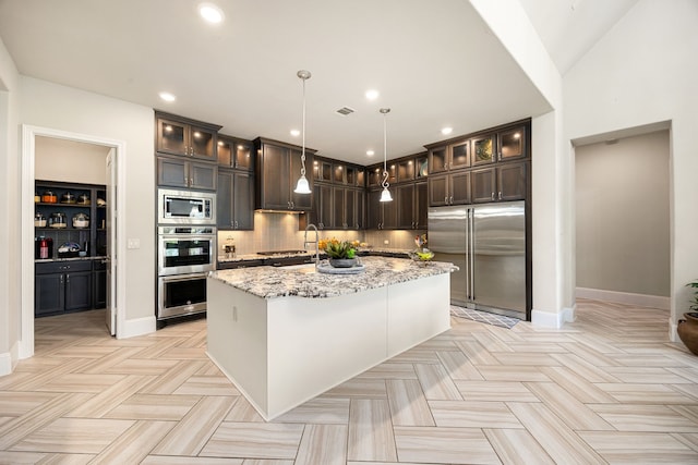 17023 Sweet Bay Ct, Conroe, TX 77385 - kitchen featuring built in appliances, dark brown cabinetry, light stone countertops, decorative backsplash, and a center island with sink