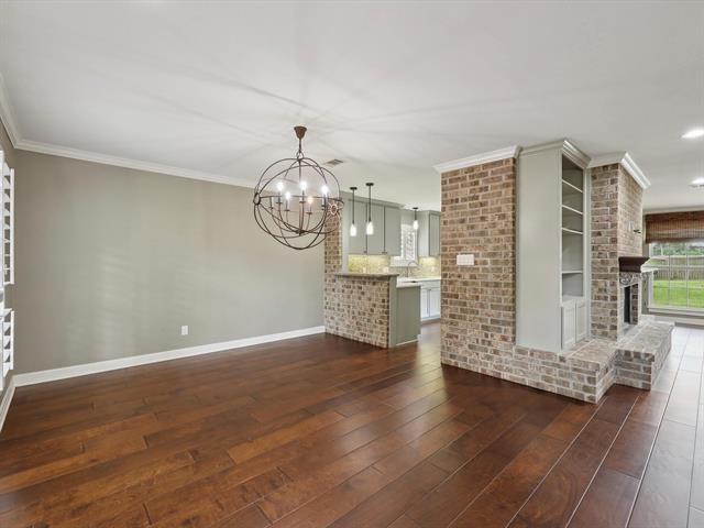 1135 Ivy Ln, Beaumont, TX 77706 - unfurnished living room with a chandelier, dark wood-style flooring, crown molding, and a brick fireplace