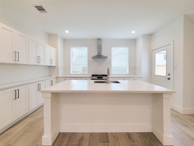 6735 Cohn St, Houston, TX 77091 - kitchen with wall chimney exhaust hood, an island with sink, light countertops, light wood-style flooring, and white cabinetry
