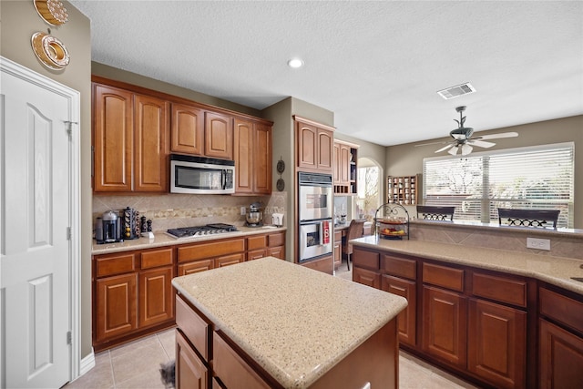 18815 Manleigh Ct, Tomball, TX 77377 - kitchen featuring appliances with stainless steel finishes, backsplash, a ceiling fan, a kitchen island, and brown cabinetry