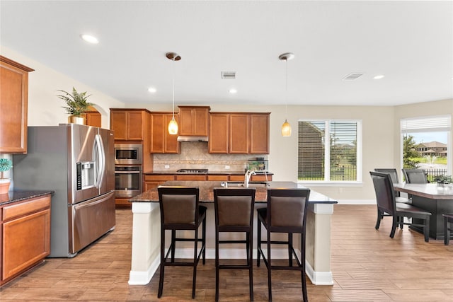 3614 Bonnie Bend Ln, Katy, TX 77494 - kitchen with appliances with stainless steel finishes, brown cabinetry, under cabinet range hood, decorative backsplash, and a kitchen bar