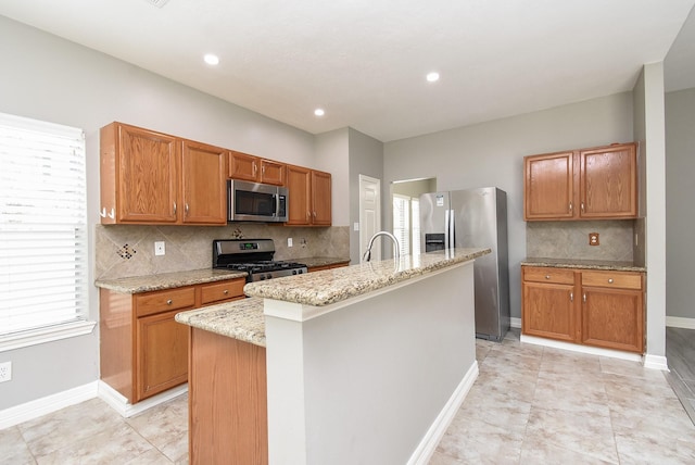 4706 Torrington Ct, Sugar Land, TX 77479 - kitchen featuring appliances with stainless steel finishes, light stone counters, a kitchen island with sink, decorative backsplash, and brown cabinetry