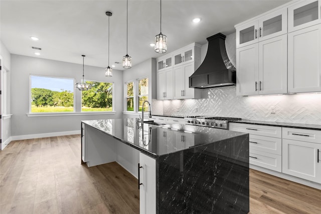 210 Hogan Ln, Hempstead, TX 77445 - kitchen featuring wall chimney range hood, light wood finished floors, decorative backsplash, a kitchen island with sink, and dark stone counters