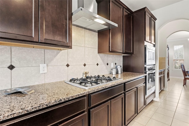 1115 Tydeman Ct, Katy, TX 77494 - kitchen with wall chimney range hood, stainless steel appliances, arched walkways, dark brown cabinetry, and light tile patterned flooring