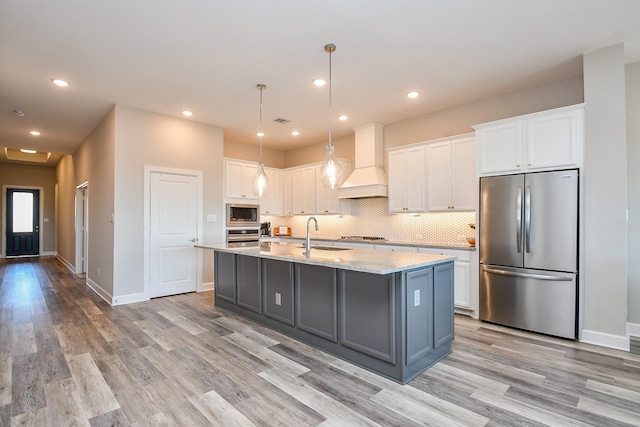 13910 Paseo De La Paz Pl, Cypress, TX 77433 - kitchen featuring appliances with stainless steel finishes, custom range hood, light wood-style floors, white cabinets, and an island with sink