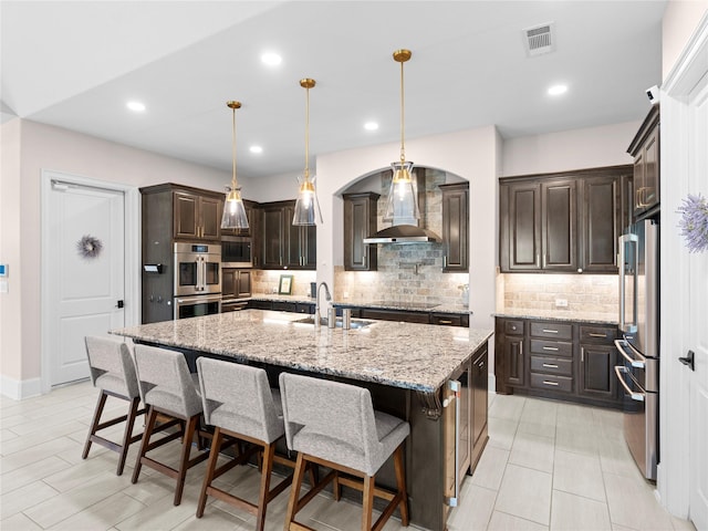 18114 Rainwater Creek Ln, Cypress, TX 77433 - kitchen with wall chimney range hood, stainless steel appliances, dark brown cabinetry, a kitchen island with sink, and decorative backsplash