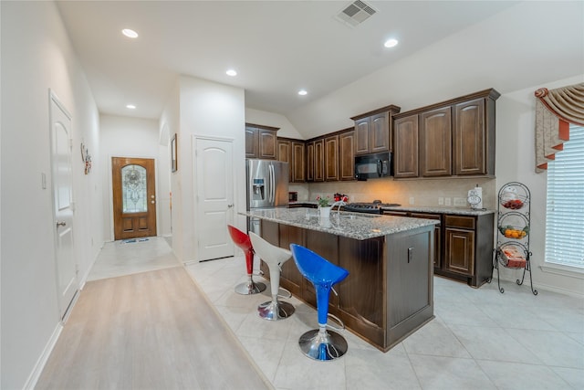 2627 Rosepoint Ct, Fresno, TX 77545 - kitchen featuring black microwave, stainless steel refrigerator with ice dispenser, dark brown cabinetry, light stone countertops, and a center island with sink