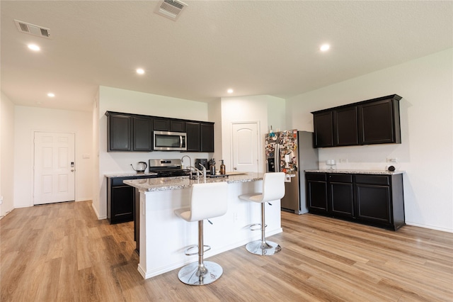 21427 Hawthorn Prairie Dr, New Caney, TX 77357 - kitchen with appliances with stainless steel finishes, light wood-style flooring, a kitchen island with sink, light stone counters, and a breakfast bar