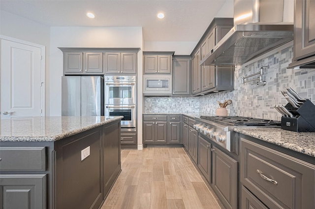 4107 Rainflower Ct, Fulshear, TX 77441 - kitchen featuring gray cabinetry, wall chimney exhaust hood, stainless steel appliances, backsplash, and light wood-type flooring