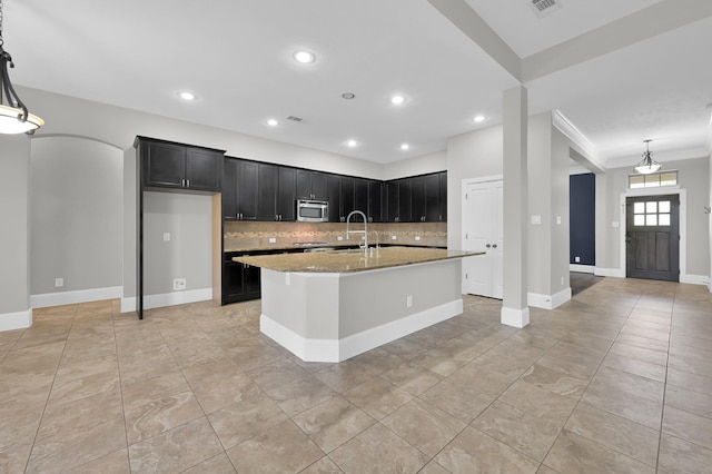 18618 Bridle Grove Ct, Tomball, TX 77377 - kitchen featuring stainless steel microwave, decorative backsplash, dark cabinets, light stone counters, and a kitchen island with sink