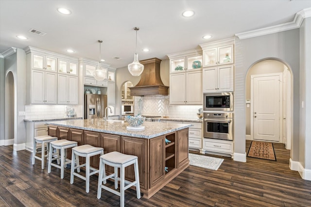32319 Tall Oaks Way, Magnolia, TX 77354 - kitchen with stainless steel appliances, arched walkways, custom range hood, a center island with sink, and dark wood-style flooring