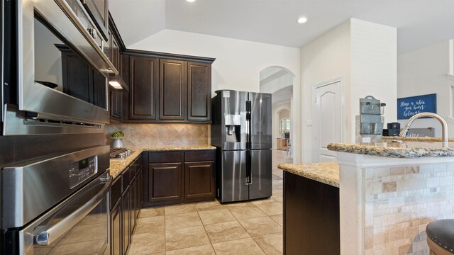 15314 Icet Creek Ave, Mont Belvieu, TX 77523 - kitchen featuring stainless steel appliances, arched walkways, light stone countertops, dark brown cabinets, and tasteful backsplash