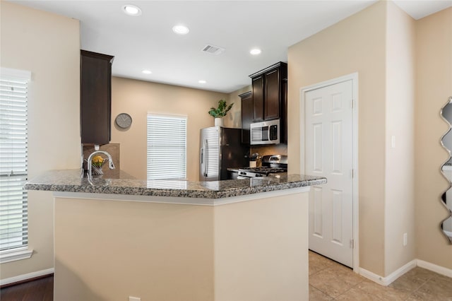 8218 Rustic Cape Dr, Cypress, TX 77433 - kitchen with stainless steel appliances, dark brown cabinets, dark stone counters, light tile patterned flooring, and a peninsula
